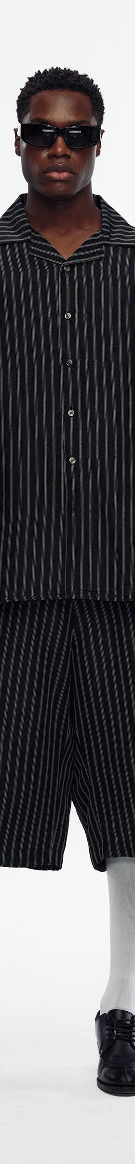 A man wearing a black pinstripe Willy Chavarria camp-collar shirt with matching wide shorts