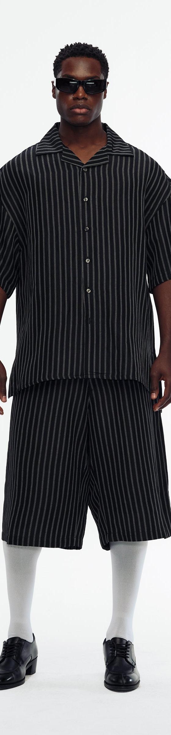 A man wearing a black pinstripe Willy Chavarria camp-collar shirt with matching wide shorts