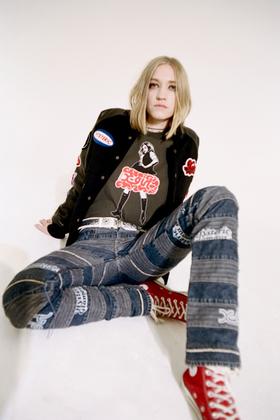 A woman wearing a black X-Girl patch varsity jacket and graphic tee with patchwork denim jeans
