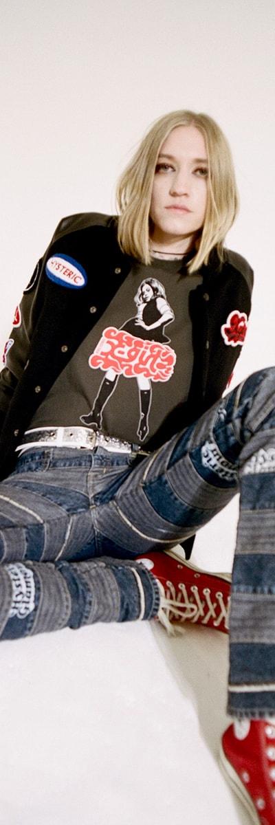 A woman wearing a black X-Girl patch varsity jacket and graphic tee with patchwork denim jeans