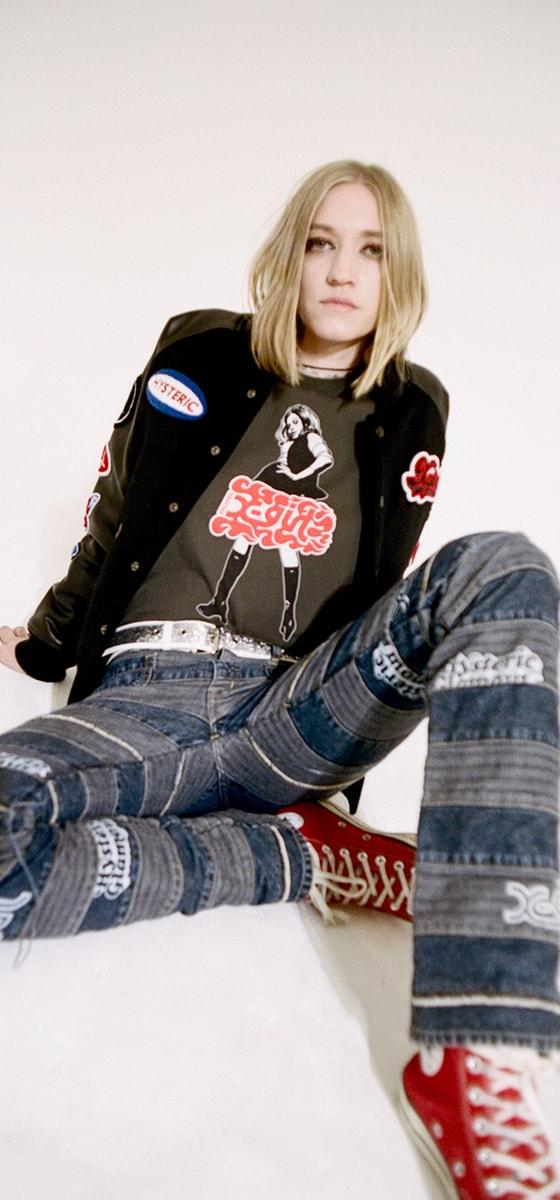 A woman wearing a black X-Girl patch varsity jacket and graphic tee with patchwork denim jeans