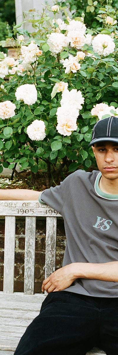 A man wearing a grey Yardsale ringer T-shirt and black cap sitting on a park bench