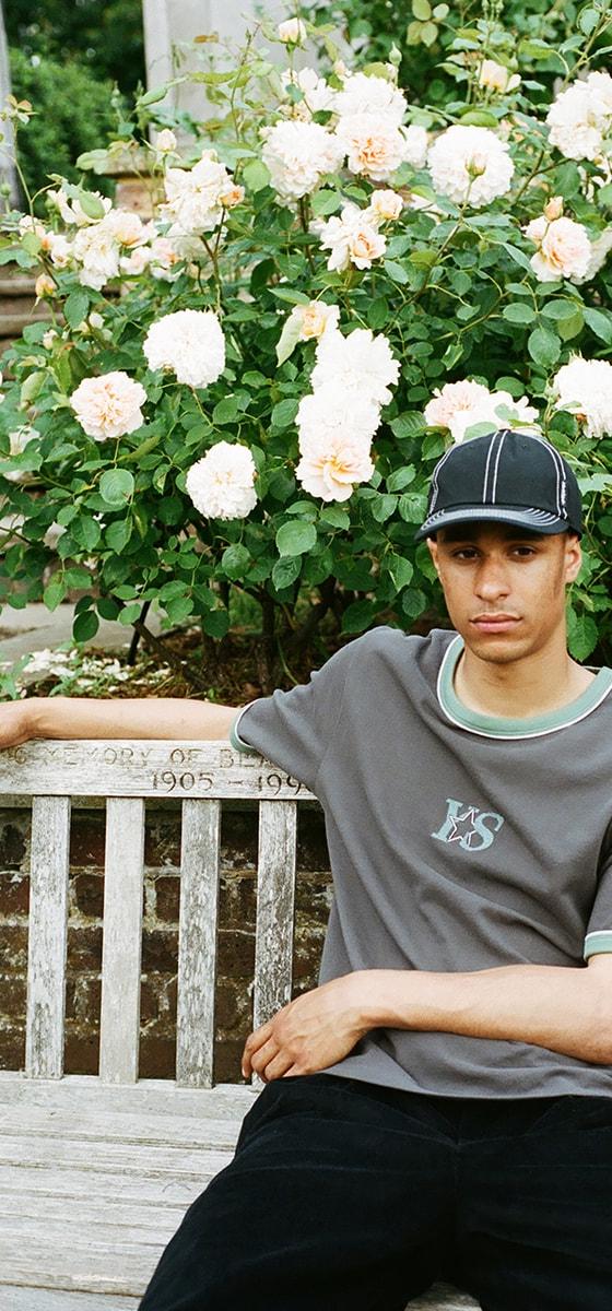 A man wearing a grey Yardsale ringer T-shirt and black cap sitting on a park bench