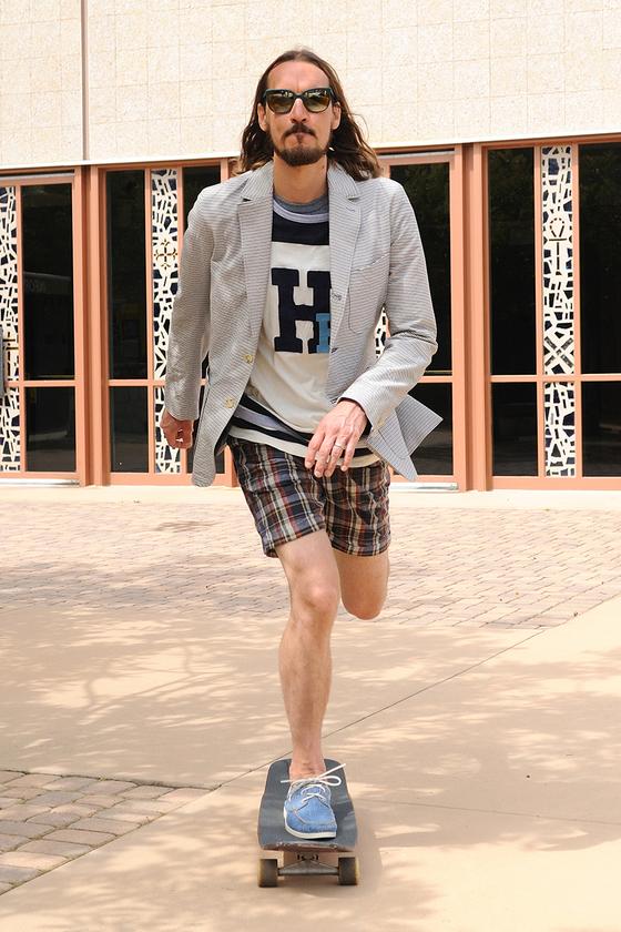 A man wearing blue Yuketen boat shoes with plaid shorts and a checked blazer, riding a skateboard
