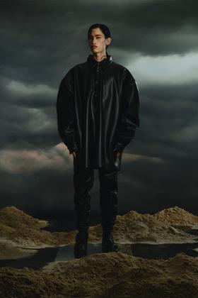 A man wearing an oversized black faux-leather YUME YUME shirt, black trousers and black boots under a storm sky