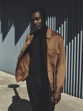 A man wearing a tan Zic suede blouson jacket over a black turtleneck and black trousers