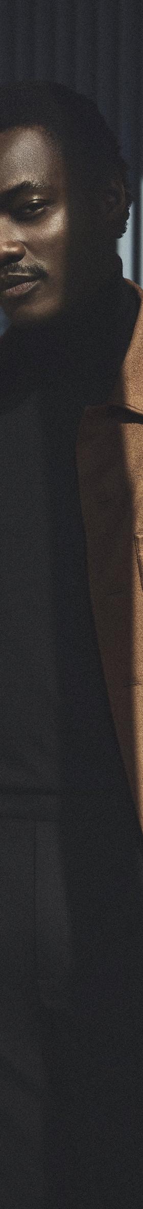 A man wearing a tan Zic suede blouson jacket over a black turtleneck and black trousers