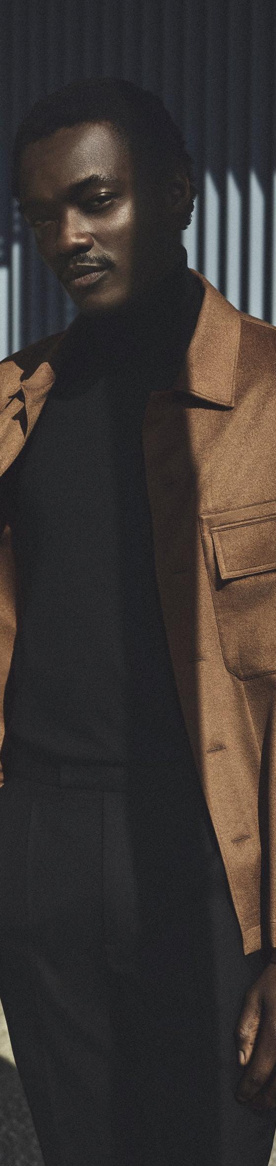 A man wearing a tan Zic suede blouson jacket over a black turtleneck and black trousers