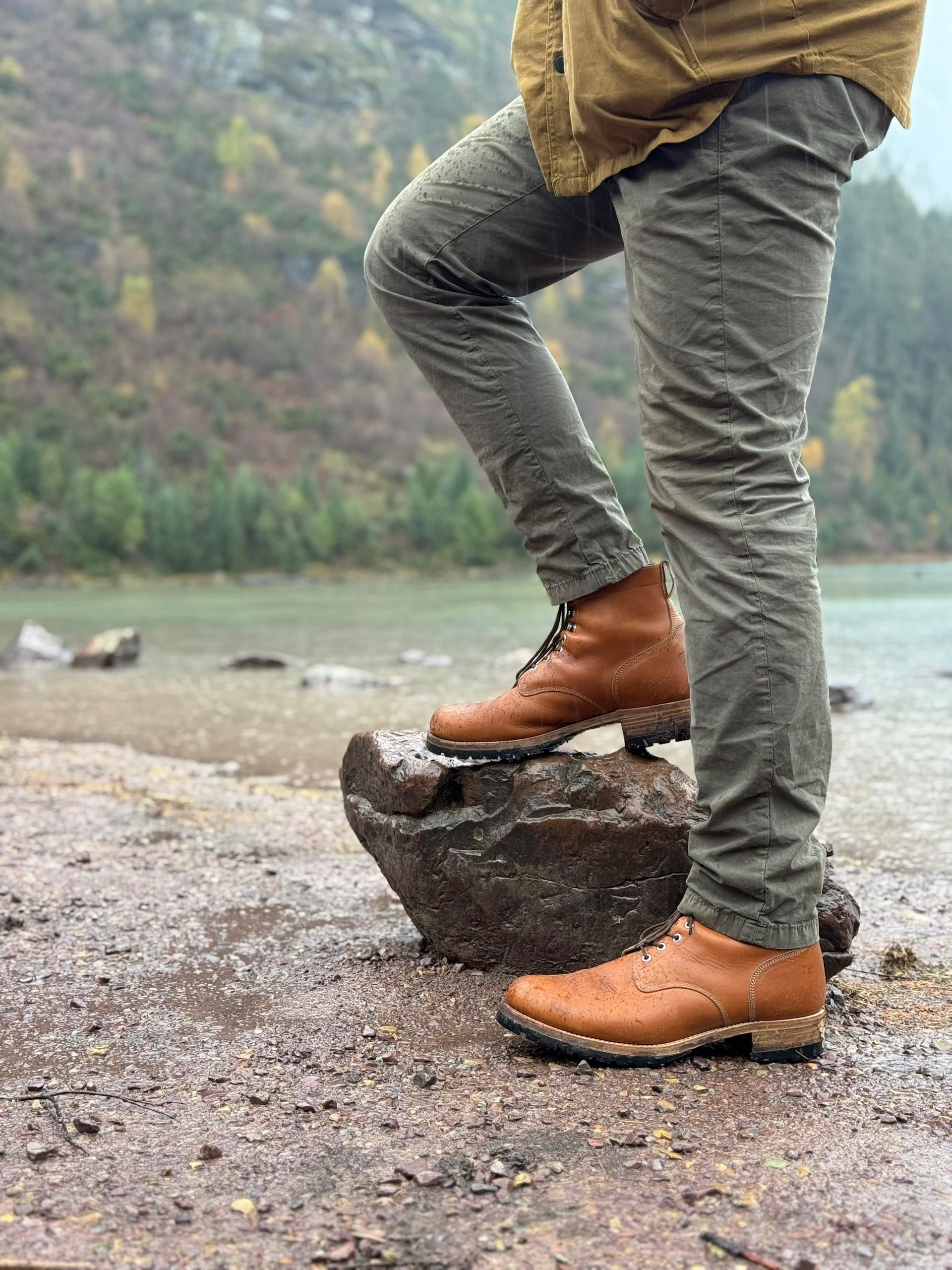 Saddleback Huntsman boots in Natural leather worn with olive trousers, one foot on a rock beside a mountain lake