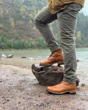 Saddleback Huntsman boots in Natural leather worn with olive trousers, one foot on a rock beside a mountain lake
