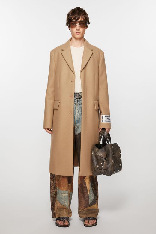 A man wearing a long camel single-breasted wool coat with an Acne Studios label patch on the cuff, worn open over a cream top, with loose printed-denim jeans pooling over dark sandals, shield sunglasses, and a studded dark-brown leather tote in one hand, against a plain light-grey background.