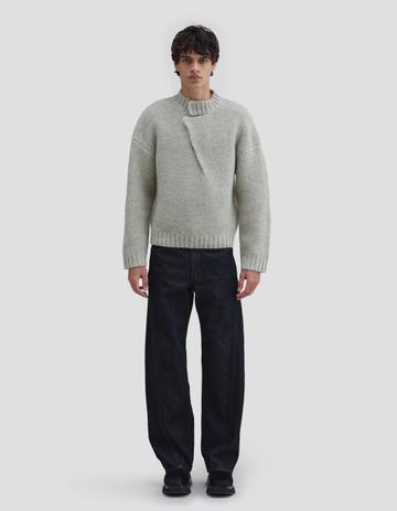A man wearing a pale grey-green chunky moss-stitch pullover with a mock neck and diagonal seam detail, dark rinse black wide curved-leg jeans and black chunky shoes, standing against a plain light-grey studio background.