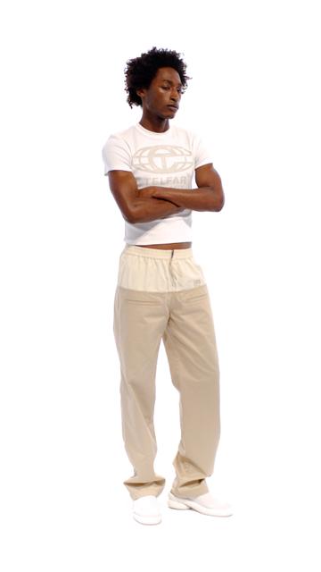 A man wearing a fitted white Telfar globe-logo T-shirt and loose two-tone khaki work pants with white slip-on shoes, standing full length with arms crossed against a plain white background.