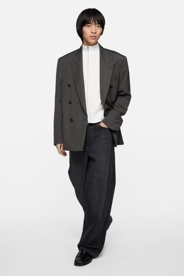 A man wearing an oversized grey pinstripe double-breasted blazer open over a white zip-neck knit, loose washed-black jeans pooling over black loafers, one hand in his pocket, against a plain white background.