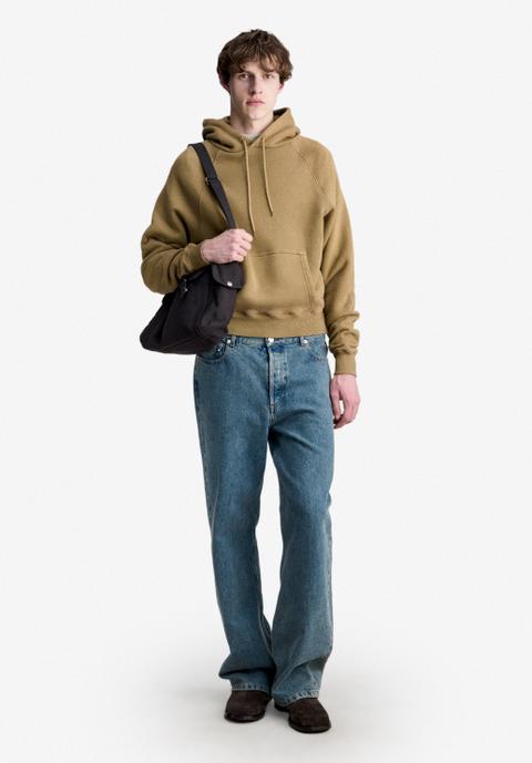 A man wearing a tan raglan pullover hoodie with a charcoal canvas shoulder bag on one shoulder, loose mid-wash straight jeans and dark-brown suede boots, standing full length on an off-white studio ground.