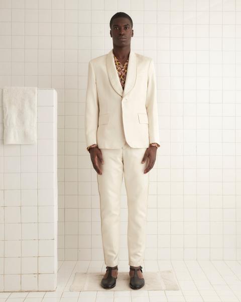 A man wearing an ecru Bode Roping shawl-collar tuxedo jacket with rope-trimmed lapels over a brown-and-yellow geometric camp-collar shirt, matching ecru trousers with rope-trimmed side seams, and black T-bar shoes, standing in a white-tiled room.