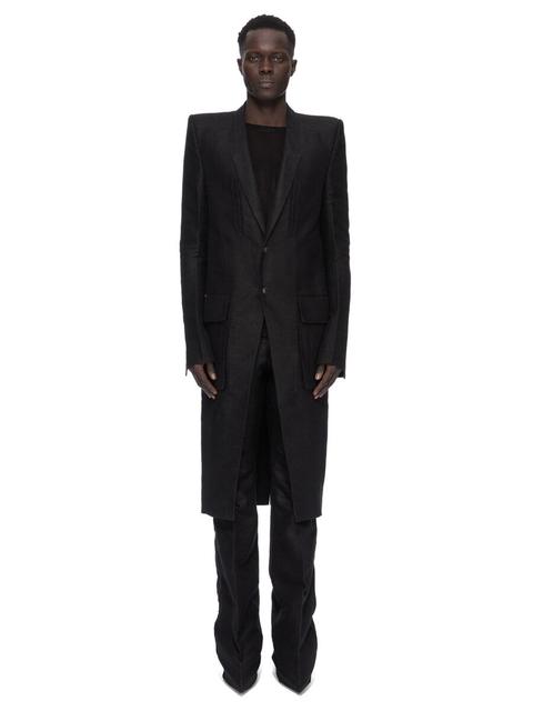 A man wearing a long, sharp-shouldered black coat buttoned at the waist over a black crew-neck top, with long black bootcut trousers stacked over pointed black shoes, against a plain white background.