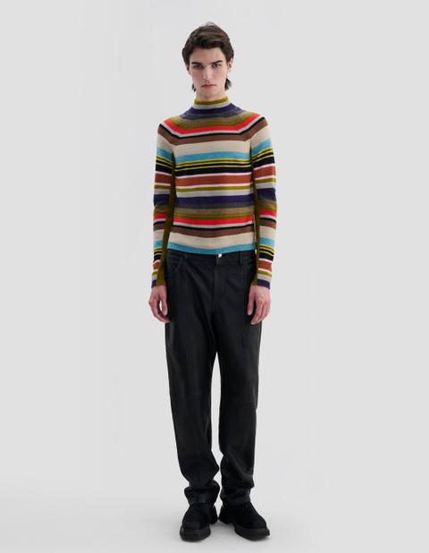 A man wearing a fitted multicolour horizontal-stripe turtleneck knit tucked over loose black leather trousers and black chunky suede shoes, standing full length against a plain light-grey studio background.