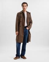 A man wearing a long brown-and-cream check coat open over an off-white ribbed henley, straight mid-blue jeans and tan pony-hair slippers, standing with one hand in his pocket against a pale-grey studio backdrop.