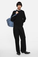 A man wearing black loose-fit jeans with a black crew-neck sweater layered over a white collared shirt, a blue suede bag on one shoulder and black western boots, standing on a plain white background.