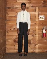 A man wearing high-waisted black pleated wide-leg trousers with turn-ups, a white bib-front tuxedo shirt with a multicolour button placket, and black bow-front slippers, standing in front of a knotty-pine wood wall.