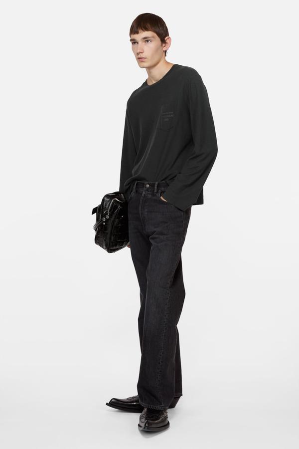 A man wearing a washed-black long-sleeve pocket tee and loose washed-black jeans over black pointed-toe leather boots, holding a black croc-effect leather bag, standing in a three-quarter turn against a plain white studio background.