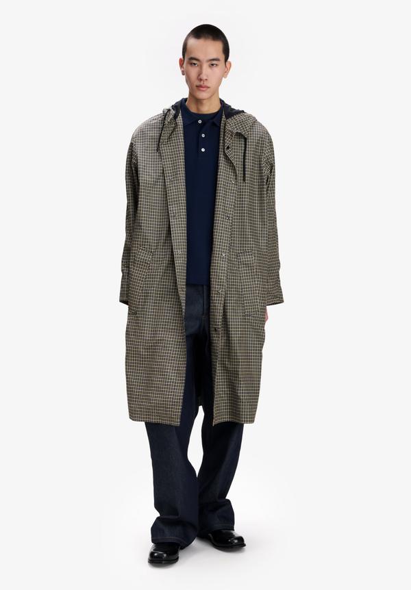 A man wearing a long hooded parka in a small navy-and-khaki check, worn open over a navy polo shirt, with wide raw-indigo jeans and black leather shoes, standing against a plain off-white background.