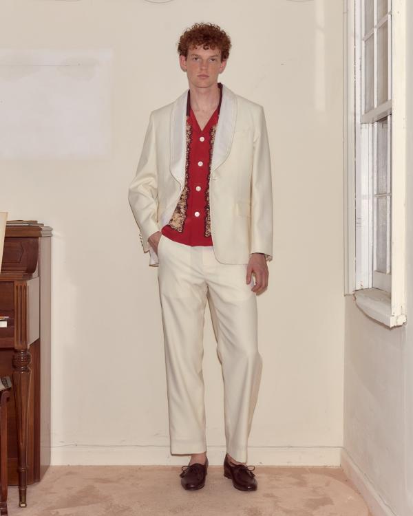 A man wearing an ivory shawl-collar tuxedo jacket with piped lapels over a red camp-collar shirt with a printed placket, matching ivory trousers and dark burgundy tassel loafers, standing in a cream-walled room between a wooden piano and a window.