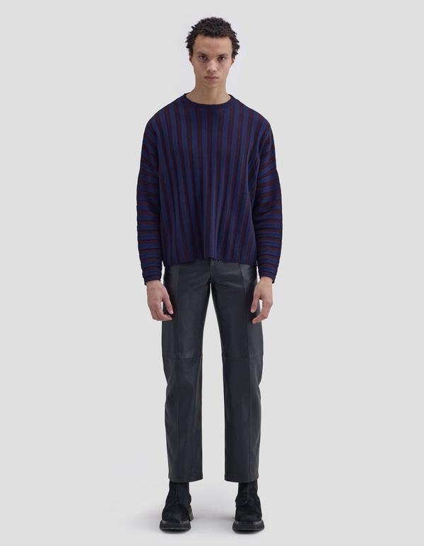 A man wearing a boxy navy-and-burgundy vertical-stripe Eckhaus Latta Keyboard sweater with straight black leather trousers and black suede chunky boots, standing full length against a plain light-grey studio background.