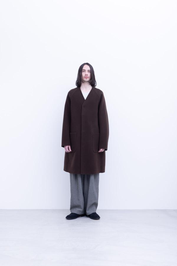 A man wearing a long dark brown collarless wool coat with patch pockets over a white top, wide grey trousers and black slip-on shoes, standing full length against a plain white background.