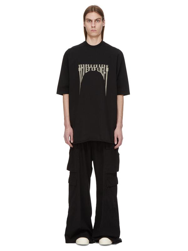 A man wearing an oversized black short-sleeve T-shirt with a cream gothic-lettering print, very wide black cargo trousers pooling over off-white sneakers, standing full length against a plain white studio background.
