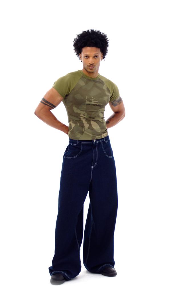 A man wearing a fitted olive camo-print raglan tee and very wide dark-indigo jeans with white contrast stitching and black shoes, standing full length with hands on his hips against a plain white background.