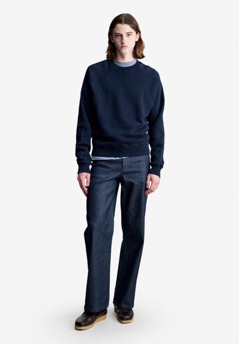A man with shoulder-length hair wearing a navy raglan crewneck sweatshirt over a pale-blue tee, dark raw-indigo straight-leg jeans and burgundy leather moccasins with crepe soles, standing against a plain off-white studio background.