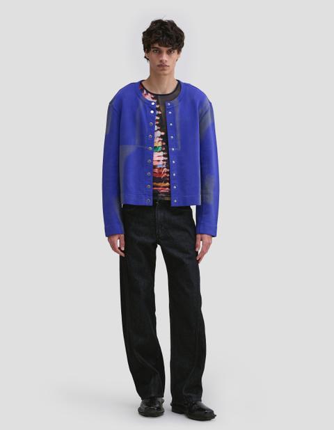 A man wearing a cobalt-blue sprayed snap-front cardigan open over a multicolour printed mesh top, black wide straight jeans and black leather shoes, standing full length against a light-grey studio background.