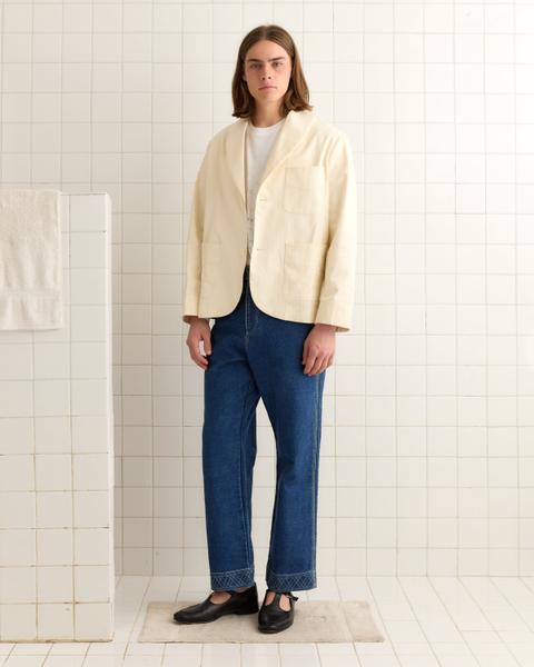 A man with shoulder-length hair wearing a cream herringbone shawl-collar smoking jacket with patch pockets over a white graphic tee, mid-blue straight-leg jeans with decorative stitched hems and black T-bar shoes, standing in a white-tiled room.