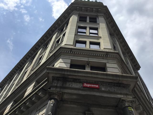 A limestone corner building shot from the sidewalk, with a red Supreme box-logo sign mounted above the ground-floor cornice