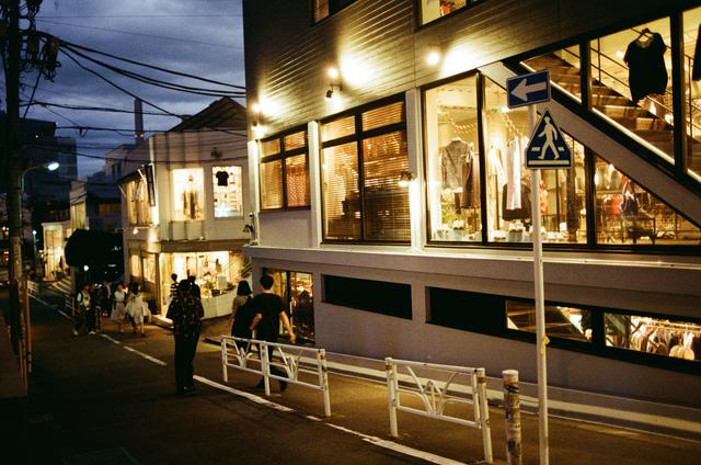 A man wearing a dark short-sleeve shirt and trousers walking past a lit second-floor boutique on a narrow Tokyo backstreet at dusk
