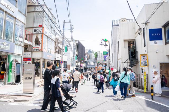 A man wearing a black short-sleeve shirt and dark trousers walking a Harajuku backstreet lined with small single-label shops and an exterior stairwell entrance, crowds of shoppers behind him