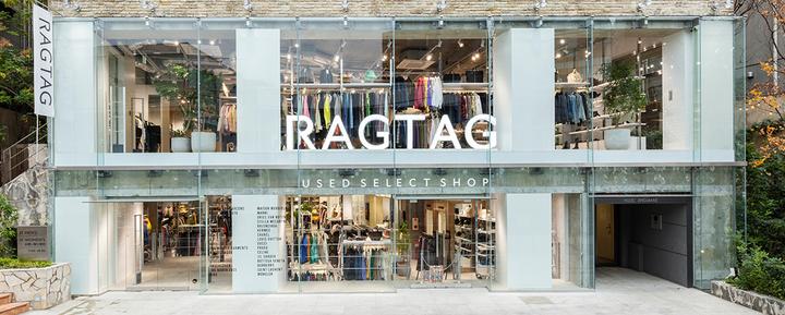 A two-storey glass shopfront with large white RAGTAG letters across the upper floor, a 'Used Select Shop' band above the entrance, a list of designer brand names on a panel beside the door, racks of clothing visible inside, and an empty paved forecourt in front, flanked by greenery and a vertical RAGTAG banner.