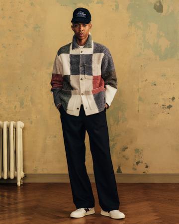 A man wearing a navy cap, a multicolour patchwork knit chore jacket over a white tee, navy wide-leg trousers and white New Balance sneakers, standing with his hands in his pockets against a weathered ochre wall beside a white radiator on a herringbone wood floor.