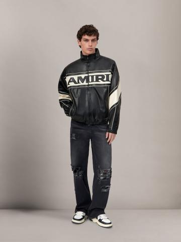 A man wearing a black leather racer jacket with a cream AMIRI logo stripe and cream sleeve panels, faded black ripped wide-leg jeans, and black-and-white sneakers, standing full length against a grey studio backdrop.