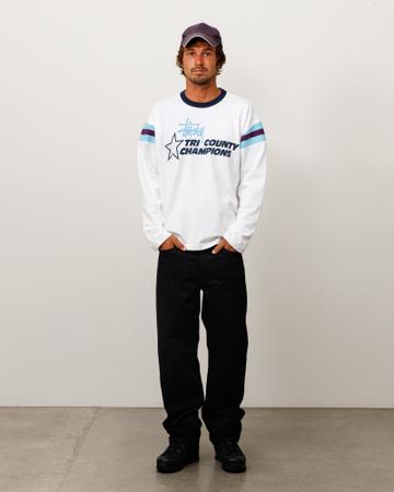 A man wearing a washed purple cap, a white long-sleeve ringer tee with a navy collar, the Stüssy script logo and a 'Tri County Champions' star graphic, light blue and maroon striped sleeves, black straight-leg jeans and black boots, standing with his hands in his pockets against a white studio wall.