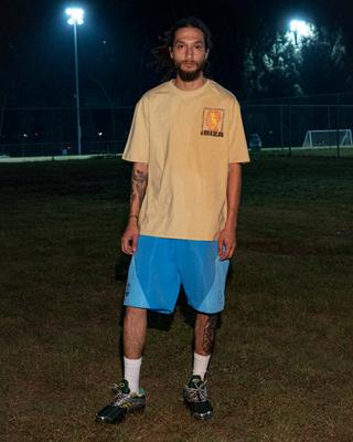 A man wearing a pale-yellow Brain Dead 'Ibiza' graphic tee, sky-blue panelled shorts, white crew socks and green-and-silver runners, standing on a floodlit football field at night with a goal in the background.