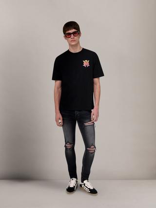 A man wearing red-tinted sunglasses, a black T-shirt with a yellow-to-pink gradient MA logo on the chest, faded black skinny jeans with ripped knees backed in pink, and black-and-white skeleton sneakers, standing full length against a grey studio backdrop.