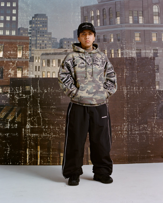 A person wearing a black cap, a camo hoodie with large black gothic-letter graphics across the chest and sleeves, baggy black track pants with white piping, and black sneakers, standing hands-in-pocket before a painted New York skyline backdrop.