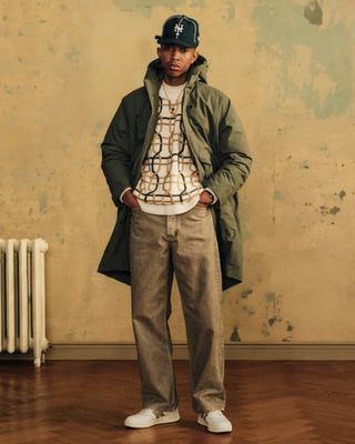 A man wearing a dark green NY Mets cap, an olive hooded parka worn open over a cream crewneck sweater with a chain-link pattern, washed khaki-brown wide-leg jeans and white retro sneakers, standing with his hands in his pockets against a weathered ochre wall beside an old radiator.