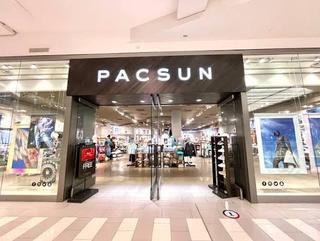 A PacSun store in a shopping mall, with illuminated white PACSUN letters on a dark wood fascia above an open glass shopfront; clothing rails, tee tables and shoe displays fill the bright interior, framed by poster windows and a striped tile floor.