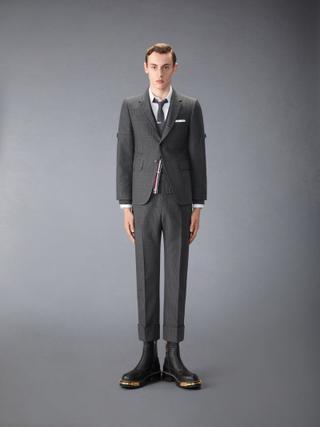 A man wearing a shrunken grey check suit with armbands, cropped cuffed trousers, a white shirt, grey tie, a cardigan with a red, white and blue striped placket, and black leather boots with gold hardware, standing against a grey studio backdrop.