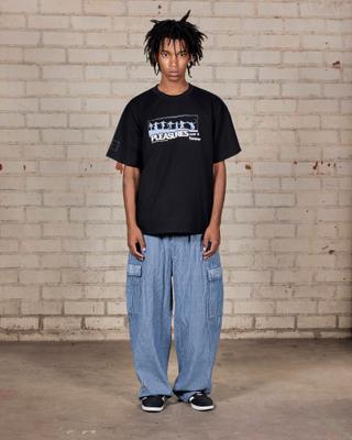 A man wearing a black PLEASURES graphic tee with a pale-blue stage-dive print, light-blue striped wide-leg cargo pants and black sneakers, standing full length against a whitewashed brick wall.