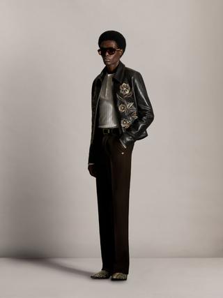 A man wearing tinted aviator sunglasses, a distressed black leather zip blouson with floral leather appliqué over a cream ribbed henley, brown pinstripe straight trousers and snakeskin boots, standing full length against a grey studio backdrop.